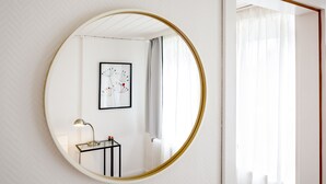A round mirror with a gold frame on a white wall, reflecting a room with a framed artwork, a small table with a lamp, and a window with sheer curtains. - Greenhouse Apartments (Helgoland)