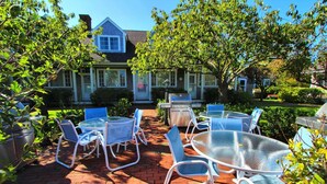 An outdoor patio with round glass tables and metal chairs, a barbecue grill, and a brick pathway leading to a house. - Brant Point Courtyard (Nantucket)