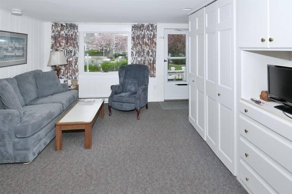 A modern living room with a grey sofa, a round armchair, and a wooden coffee table. There is a TV on a white cabinet, and a window with curtains.
