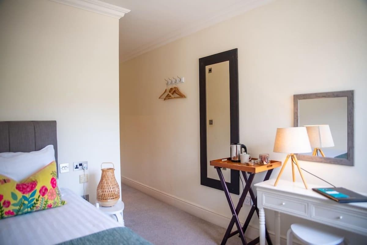 Quarto de hotel com cama, mesa de cabeceira, luminária, espelho e pequena mesa com chaleira e xícara.