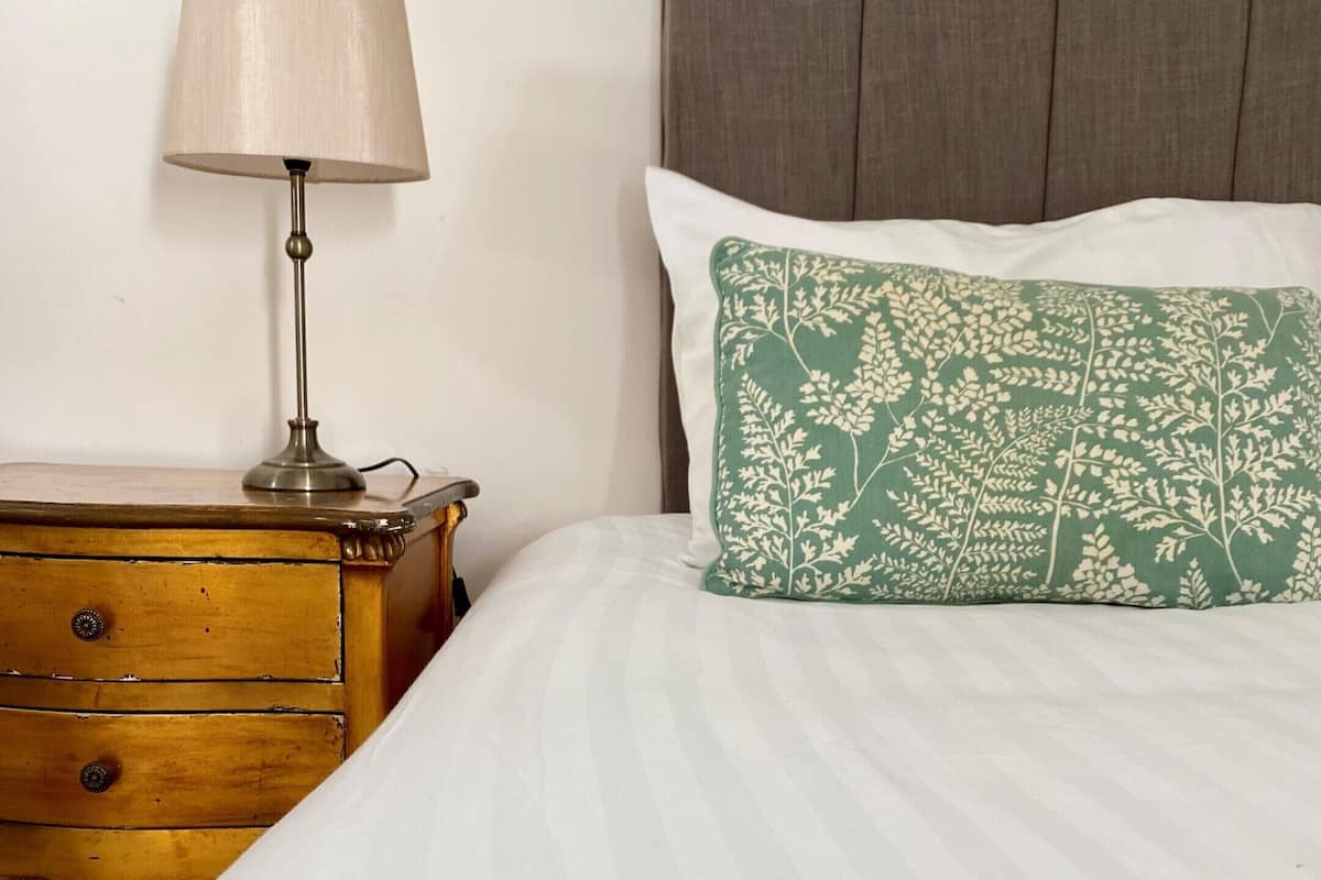 Ein ordentlich bezogenes Bett mit einer grünen Decke, einem Kissen mit Blättermotiv, einer Lampe auf einem hölzernen Nachttisch und einem grauen, gepolsterten Kopfteil.