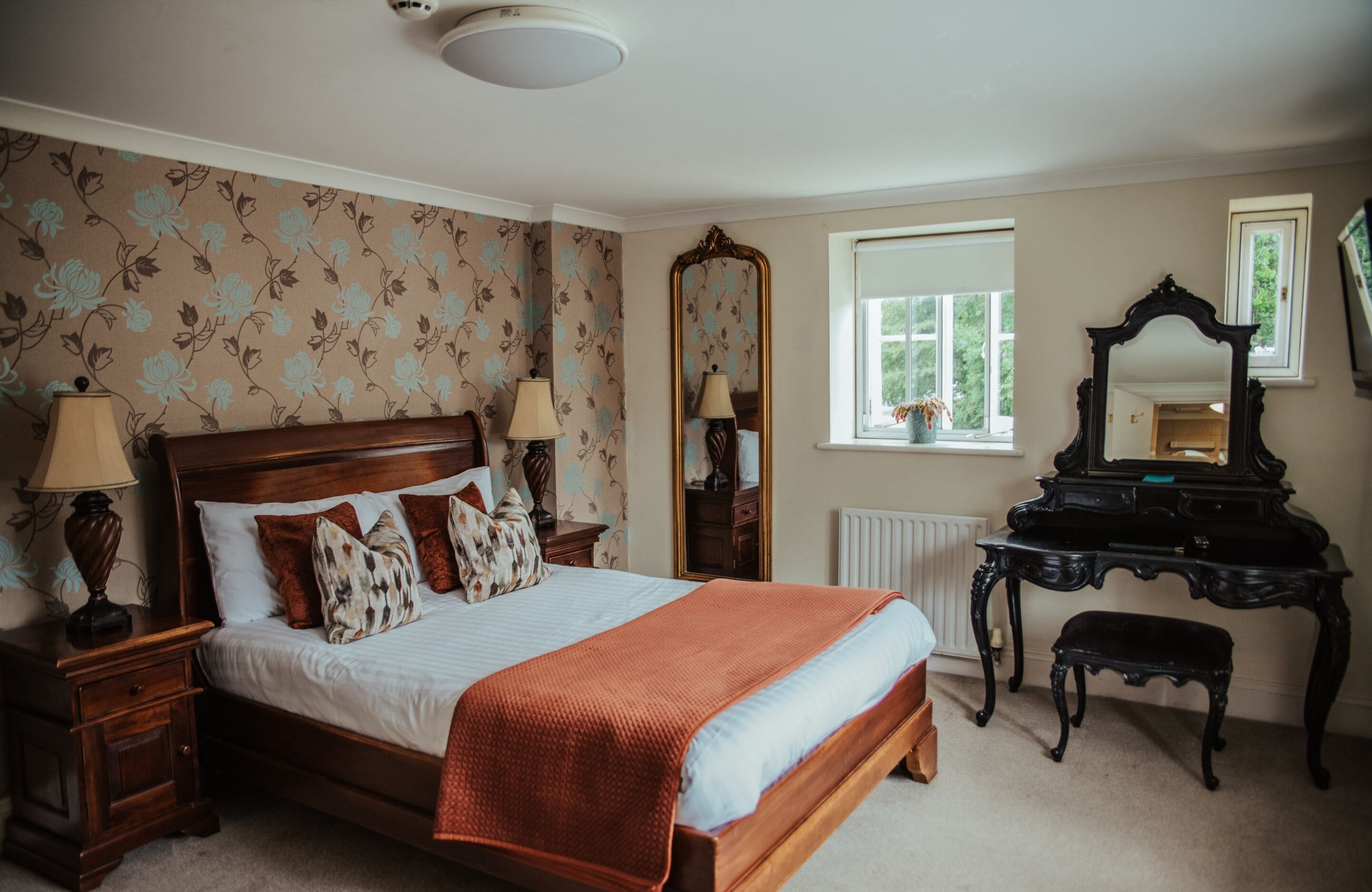 A bedroom with a large bed, two bedside tables with lamps, a dresser, a mirror, and a window with a view of greenery.