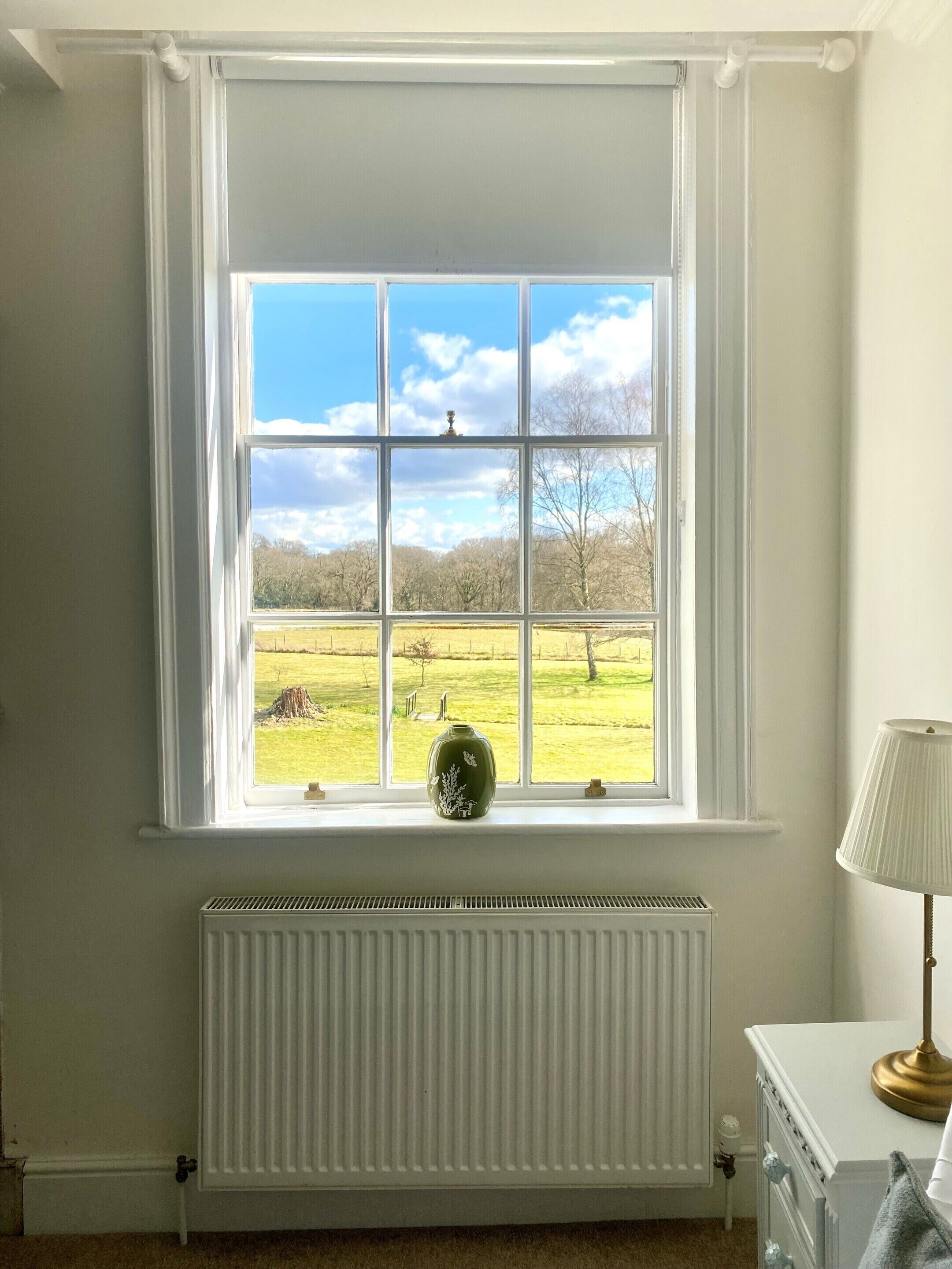 Une pièce avec une grande fenêtre, un radiateur en dessous et une lampe sur une commode.