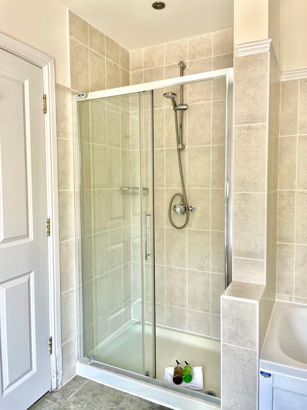 A glass-enclosed shower with a handheld showerhead and a wall-mounted faucet, a white door, and a tiled floor.