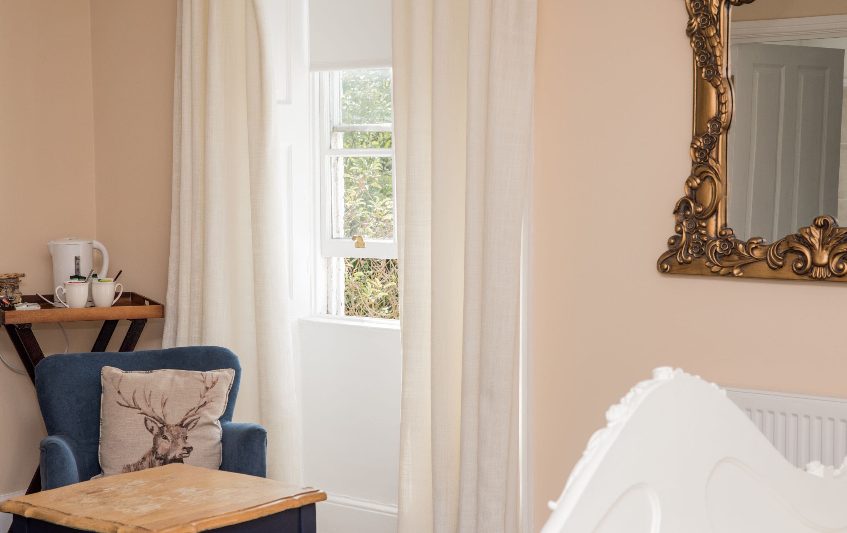 A room with a blue chair, a wooden table, a white radiator, and a large ornate mirror on the wall.