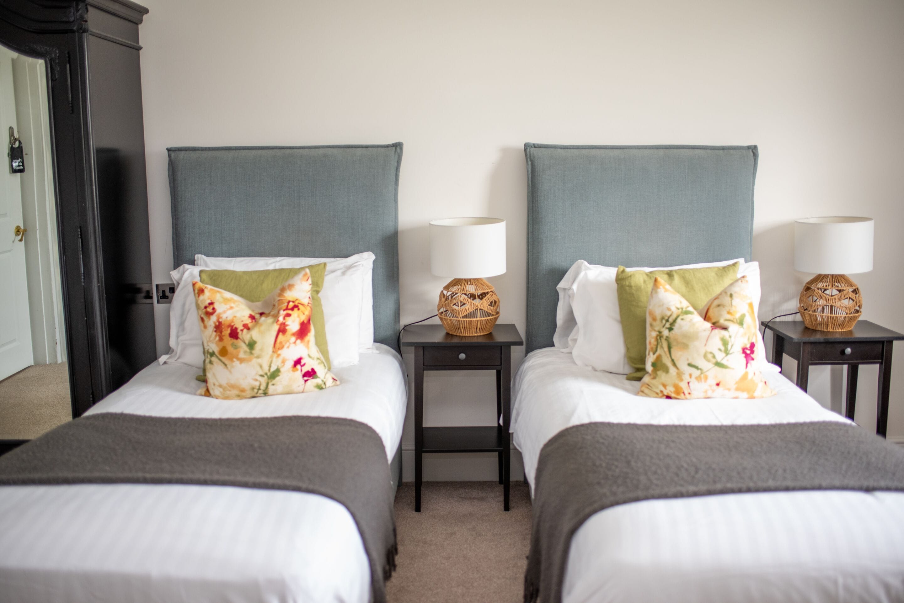 A hotel room with two beds, each with a grey headboard, white bedding, and floral-patterned pillows. There are two bedside tables with lamps and baskets.