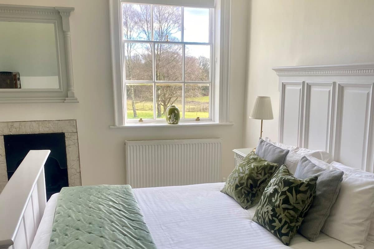 Ein Schlafzimmer mit Bett, Fenster mit Blick, Heizkörper und Spiegel an der Wand.