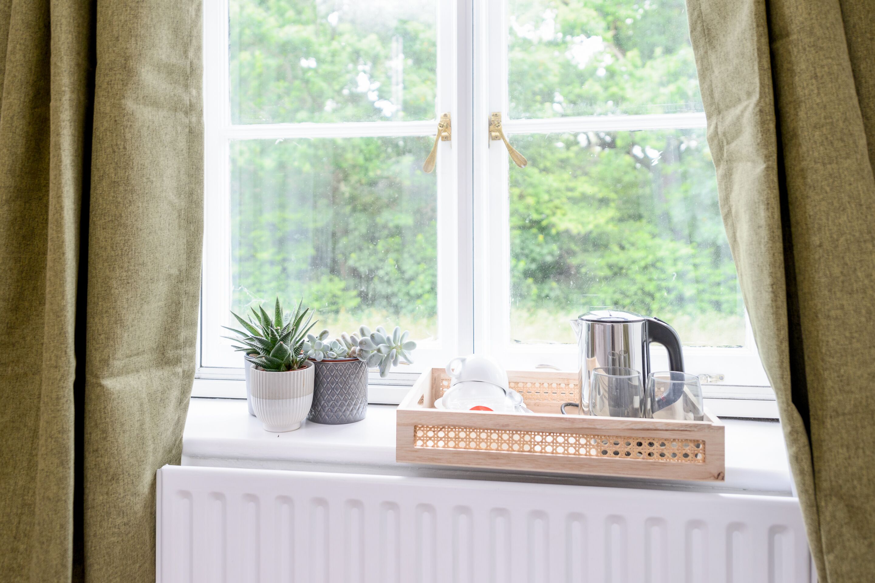 A window with a view of greenery, a radiator, and a tray with a kettle and glasses.