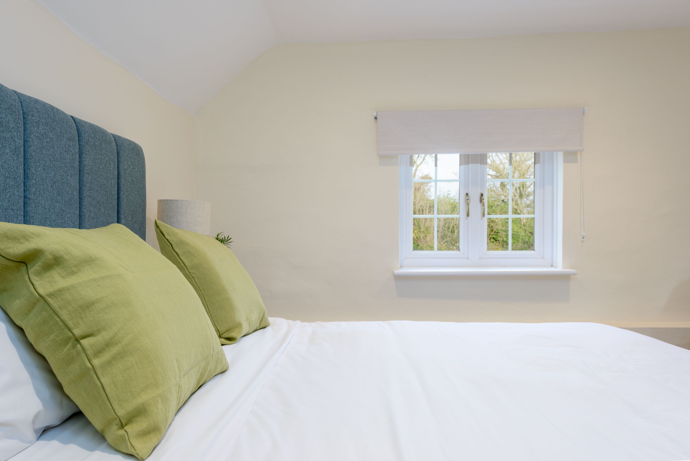 Un dormitorio con cama, almohadas, cabecera, ventana con persianas y lámpara.