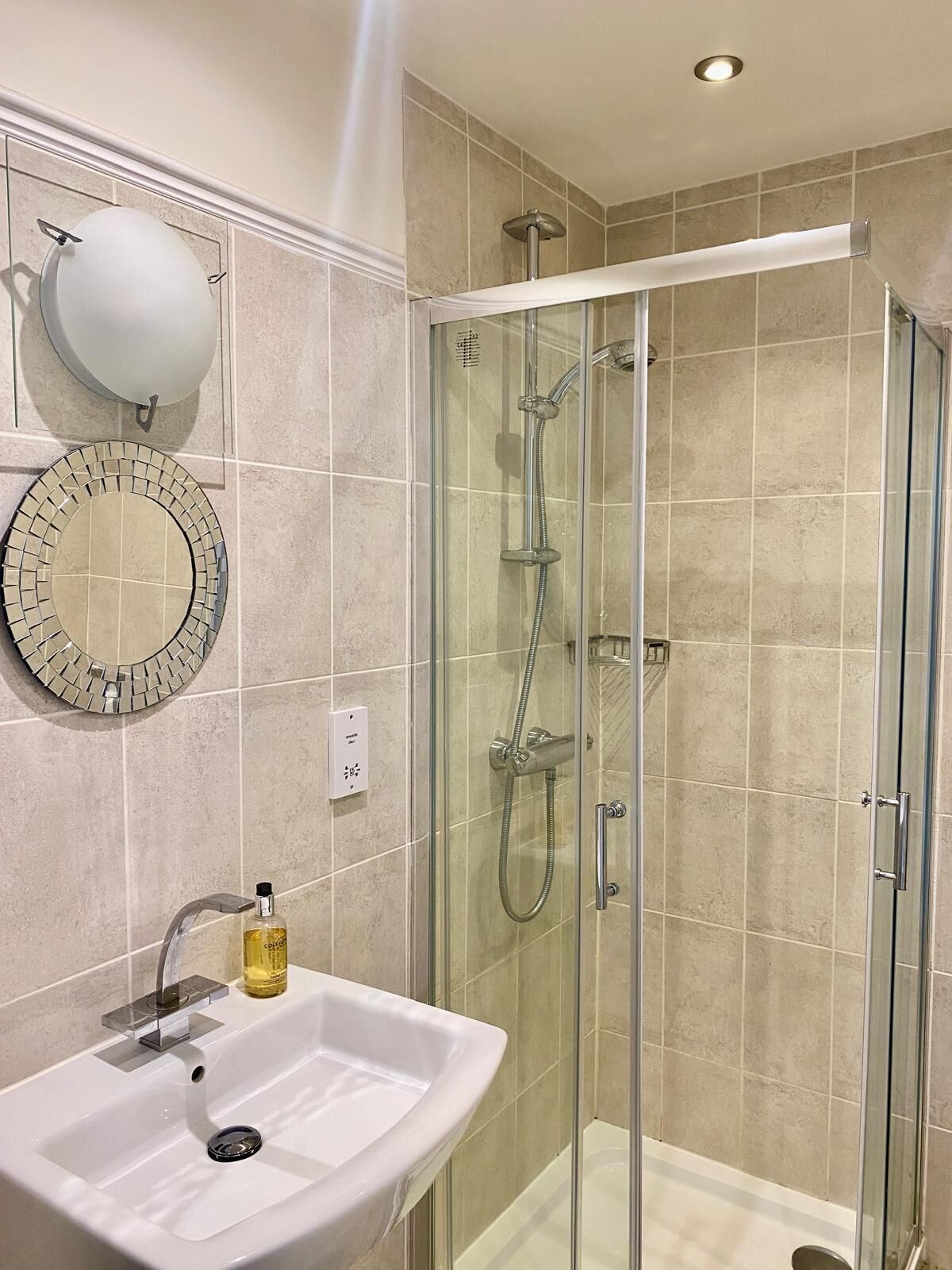 Un baño con ducha de vidrio, lavamanos con espejo y un aplique de pared.