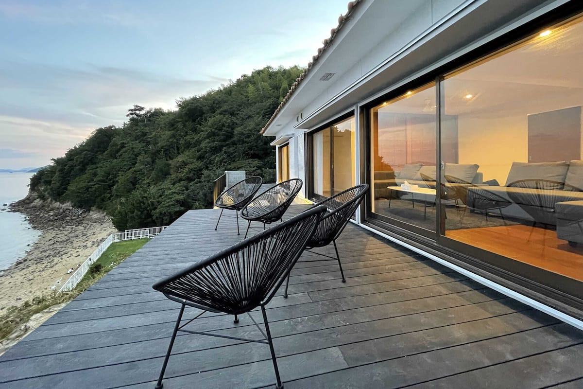 Um terraço moderno ao ar livre com duas cadeiras pretas e uma mesa, com vista para uma praia e um morrobo arborizado.
