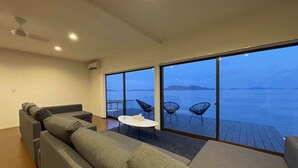 Un salon moderne avec un grand canapé, une petite table ronde et deux fauteuils. Il est doté d’une grande porte-fenêtre coulissante qui donne sur un balcon offrant une vue sur la mer et les îles lointaines.