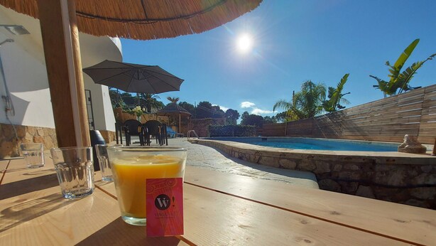 Une table en bois avec un verre de jus d’orange et un verre d’eau, donnant sur une piscine et un espace de détente extérieur.