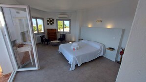 Une chambre à coucher avec un lit, deux chaises, une petite table et une vue sur l’extérieur grâce à de grandes fenêtres.