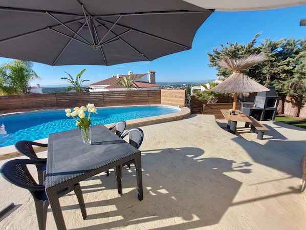 A poolside area with a table and chairs, a large umbrella, and a barbecue grill. - Modern villa with stunning views and swimming pool. (L'Escala)