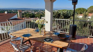 Une terrasse avec une table dressée pour le repas, des chaises, un vase rempli de fleurs et une vue sur la ville et les montagnes.