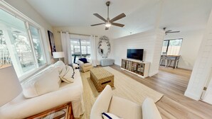 A spacious living room with a white sofa, a coffee table, and a TV on a wooden cabinet. There is a ceiling fan, a large mirror, and a view of the outdoors through the windows. - Relaxing Renovated Cottage on  Lake June in Winter with Dock! (Lake Placid)