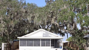A white single-story house with a covered porch, a small deck, and a dock extending into a body of water. - Relaxing Renovated Cottage on  Lake June in Winter with Dock! (Lake Placid)