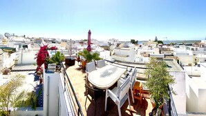 A rooftop terrace with a table and chairs, surrounded by potted plants and a view of the cityscape.