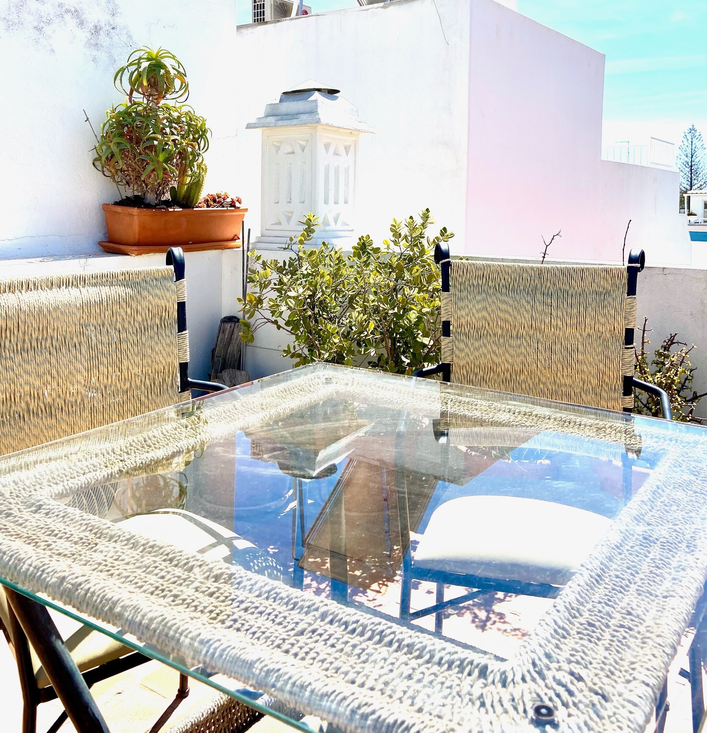 A glass-top table with woven chairs, a potted plant, and a white building in the background.