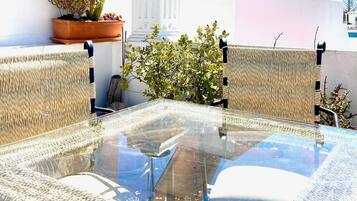 A glass-top table with woven chairs, a potted plant, and a white building in the background.