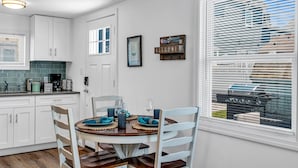 Een keuken met witte kastjes, een ronde eettafel met blauwe placematen, en een raam met jaloezieën.