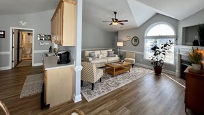 A spacious living room with a high ceiling, a dining area with wooden cabinets, and a large window providing natural light. - New! Charming Townhome 3 Minutes to Campbell (Lillington)
