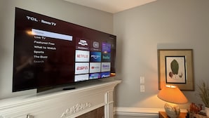 A flat-screen TV mounted on a wall above a white mantelpiece with a framed botanical print. - New! Charming Townhome 3 Minutes to Campbell (Lillington)