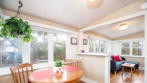 A cozy dining area with a wooden table and chairs, a hanging plant, and a view through large windows.