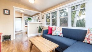 A cozy living room with a wooden coffee table, a blue sofa with colorful cushions, and large windows offering a view of the outdoors.