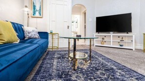 Una sala de estar con un sofá azul, una mesa de centro de vidrio, un televisor sobre un soporte blanco y una alfombra estampada.