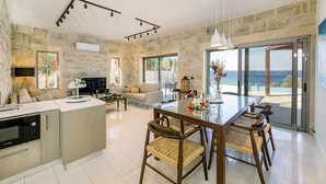 A spacious living area with a stone wall, a dining table with chairs, a kitchen area, and a large window offering a view. - Villa Corallium in Sfinari - 5 min from the beach (Sfinari)