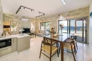 A spacious living area with a stone wall, a dining table with chairs, a kitchen area, and a large window offering a view. - Villa Corallium in Sfinari - 5 min from the beach (Sfinari)
