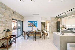 A modern kitchen with a stone wall, a dining area with a glass table, and a kitchen island. - Villa Corallium in Sfinari - 5 min from the beach (Sfinari)
