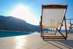 A beach chair with a mesh backrest and wooden frame, positioned on a tiled deck with a view of the ocean and mountains in the background.