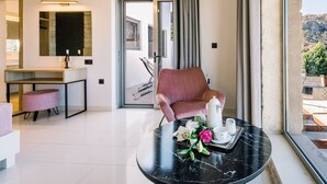 A modern hotel room with a marble table, a pink armchair, a dining area with a white countertop, and a large window with a view of the outdoors. - Villa Corallium in Sfinari - 5 min from the beach (Sfinari)