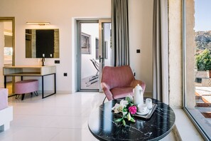A modern hotel room with a marble table, a pink armchair, a dining area with a white countertop, and a large window with a view of the outdoors. - Villa Corallium in Sfinari - 5 min from the beach (Sfinari)