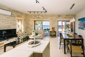 A modern living room with a stone wall, a flat-screen TV, a dining area with a table and chairs, and a view of the sea through large windows. - Villa Corallium in Sfinari - 5 min from the beach (Sfinari)