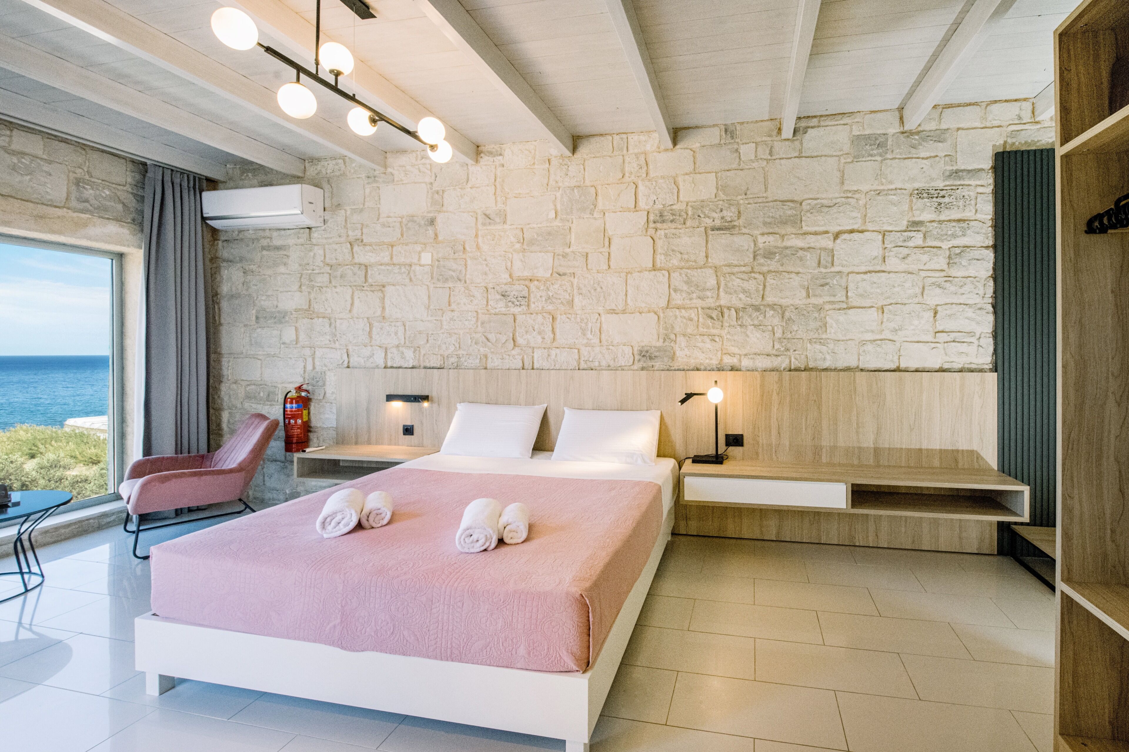 Ein modernes Hotelzimmer mit Steinwand, einem Bett mit rosa Decke, einem Nachttisch, einem Sessel und Meerblick.