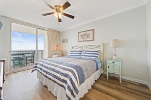 A bedroom with a large bed, a ceiling fan, a nightstand with a lamp, a framed picture on the wall, and a balcony with a table and chairs. - Beachfront Condo with On-Site Dining & Spa (Gulf Shores)