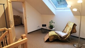 A loft bedroom with a sloped ceiling, a skylight, a wooden staircase, a cushioned chair, a floor lamp, and a small table with a plant.