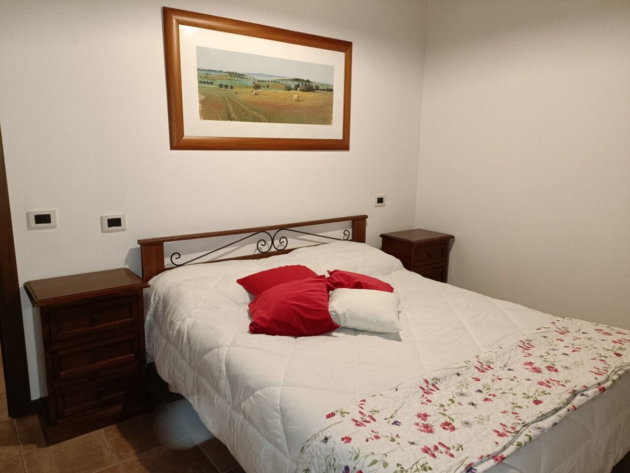 Una camera da letto con un letto, un comodino, una cassettiera, un quadro appeso al muro e una coperta con motivi floreali.