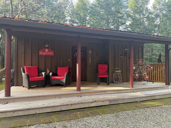 A covered outdoor seating area with wicker furniture and red cushions, a sign reading "Cousine Cabin," and a wooden deck. - Cowgirl Cabin • 2 Queens • AC • W/D • Wi-Fi (Selma)