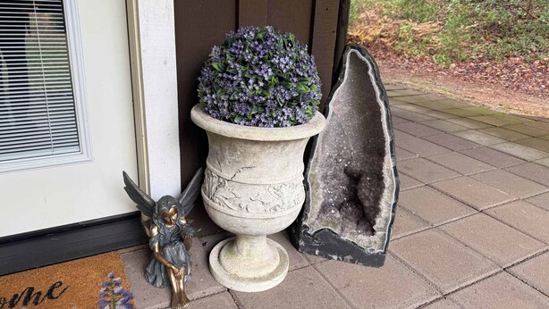 Una jardinera de piedra con flores moradas, un ángel decorativo y una escultura rocosa única en un porche con baldosas.