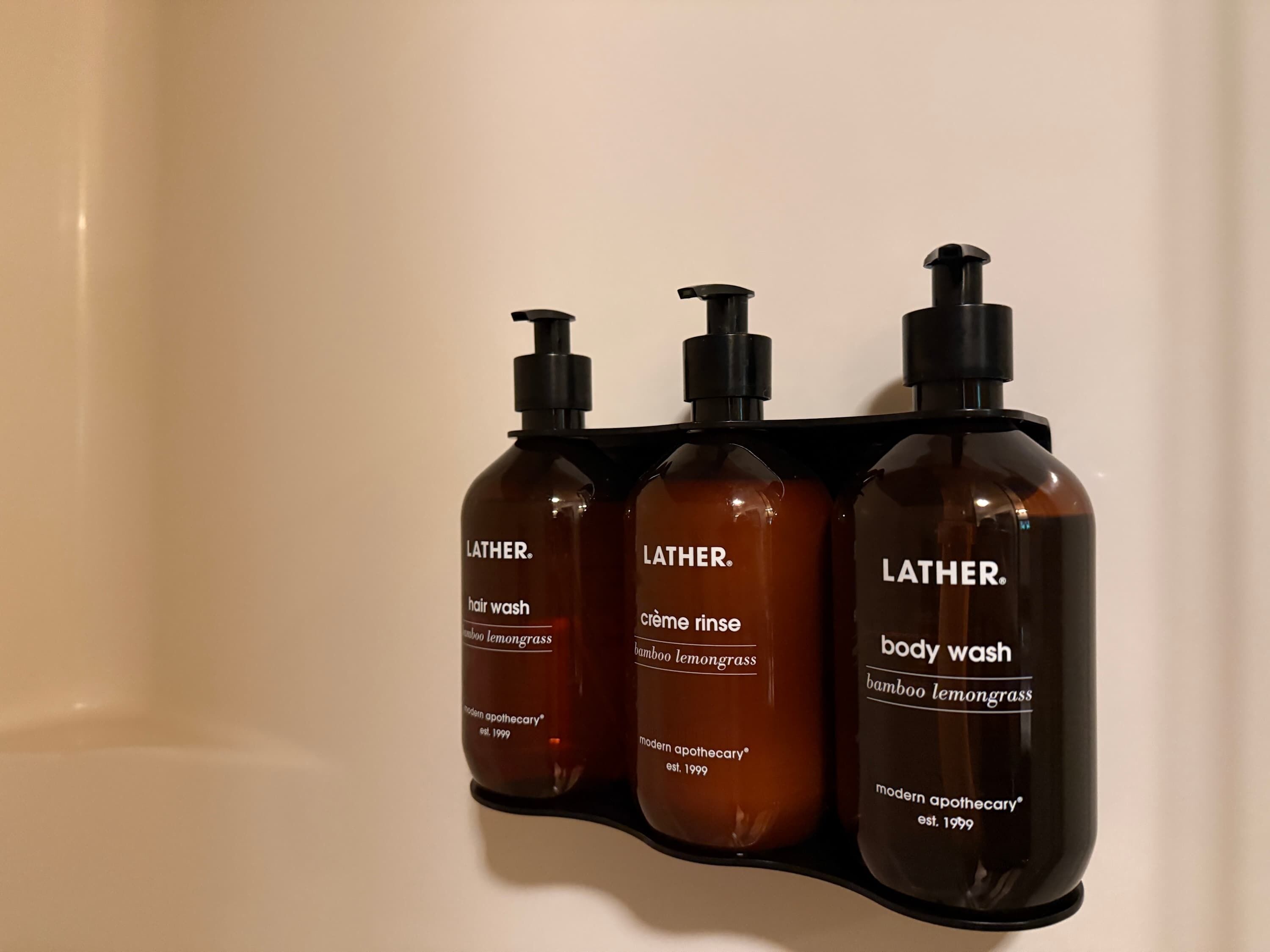 Três frascos de banho líquido Lather em um suporte preto numa parede de banheiro.