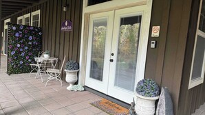 Entrada de hotel con una puerta blanca, felpudo y mobiliario exterior con sillas y macetas.