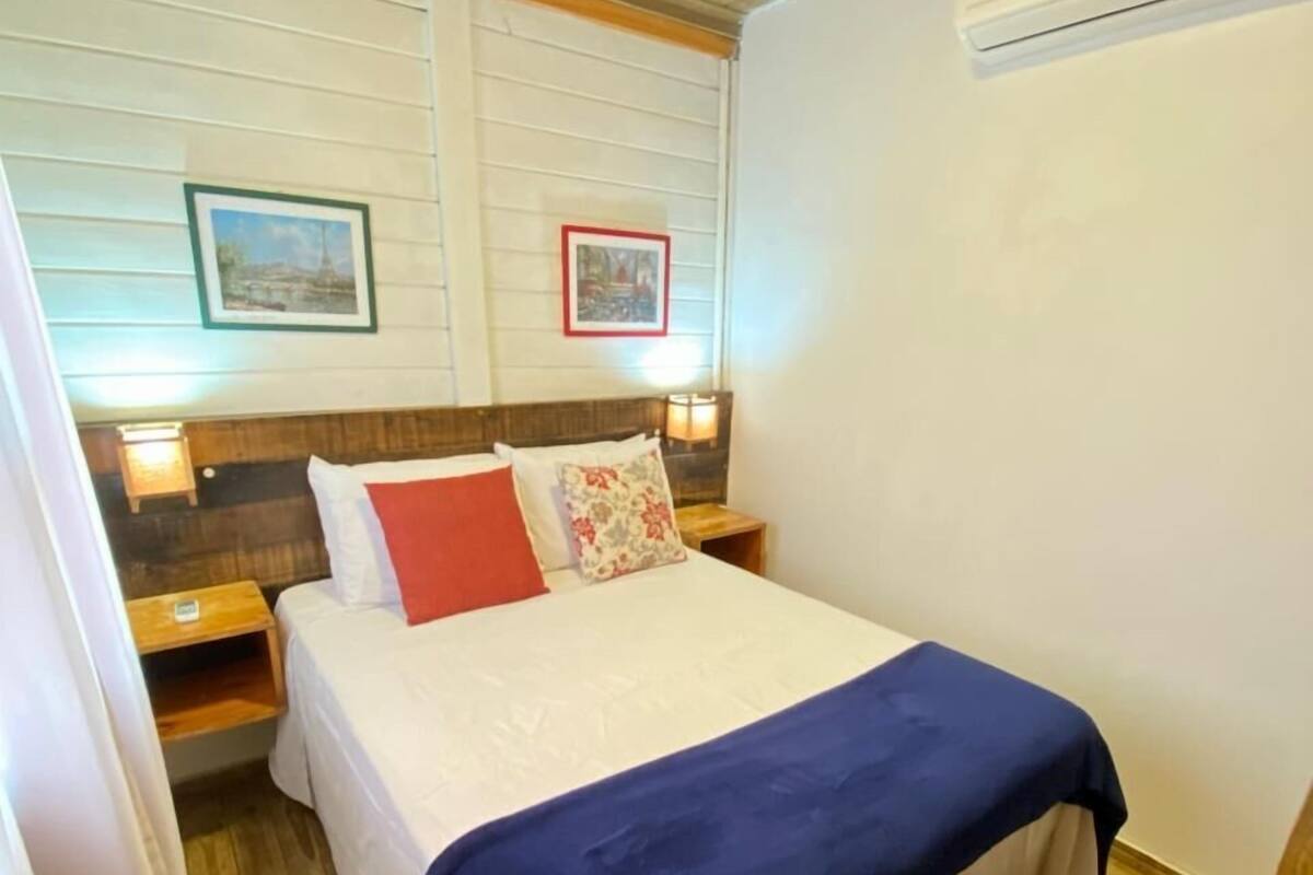A bedroom with a wooden ceiling, a bed with a blue blanket, two bedside tables with lamps, and two framed pictures on the wall.