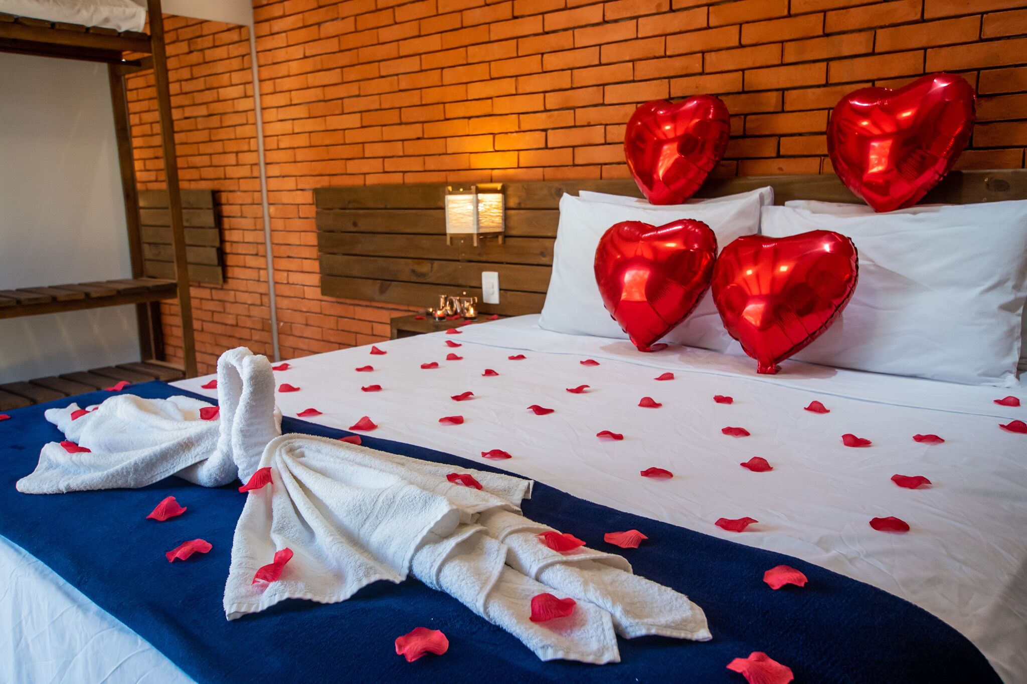 A hotel room with a brick wall, a wooden headboard, and a bed with white linens and red heart-shaped pillows.