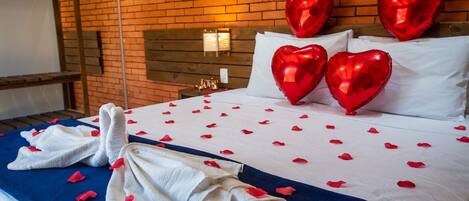 A hotel room with a brick wall, a wooden headboard, and a bed with white linens and red heart-shaped pillows.