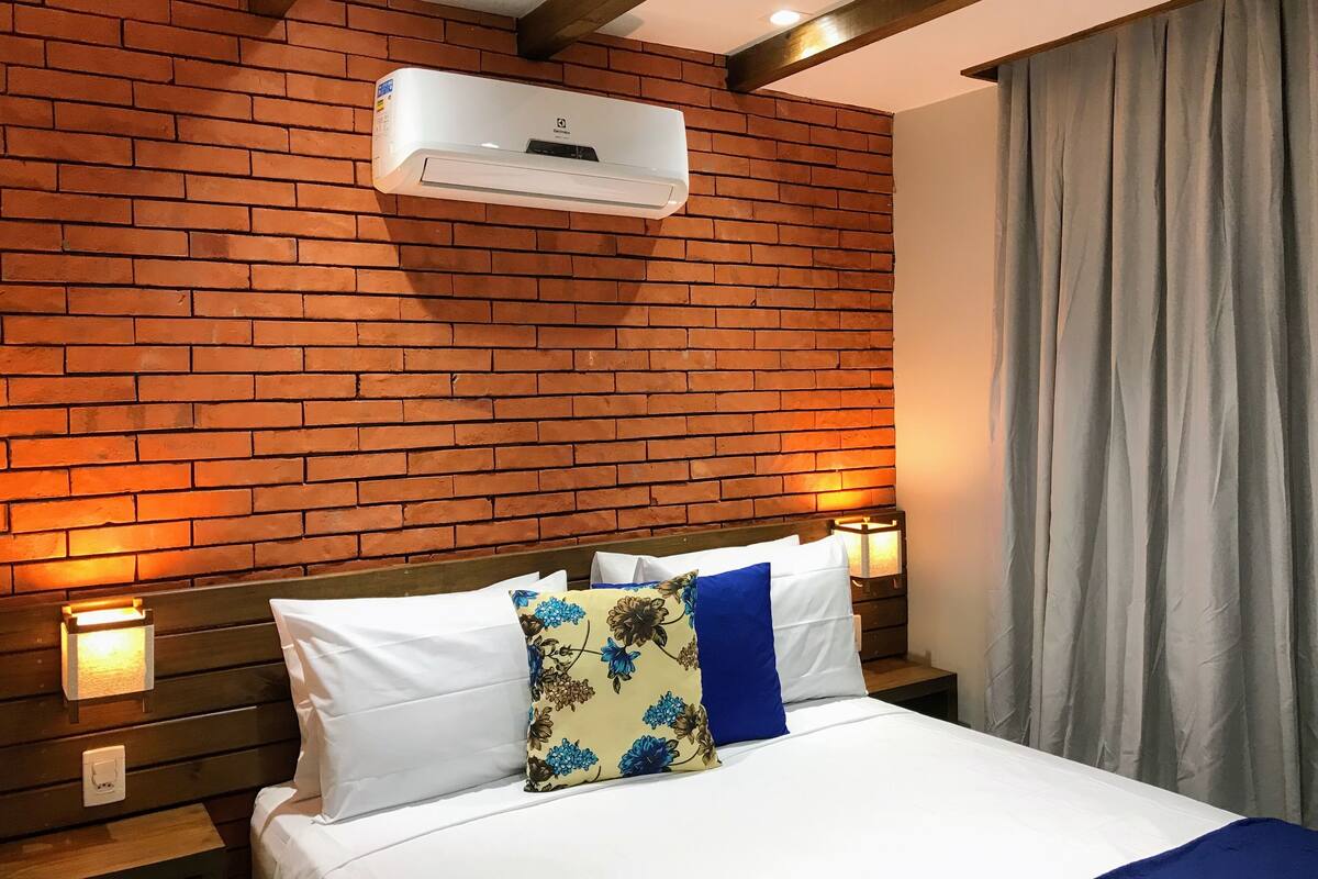 A hotel room with a brick wall, a bed with white bedding, a floral pillow, and bedside lamps.